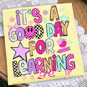 May include: A light yellow t-shirt featuring the colorful text IT'S A GOOD DAY FOR LEARNING. The design includes a pink smiley face, a red apple, a pencil, stars, flowers, and bows in various patterns like checkers, stripes, and polka dots.