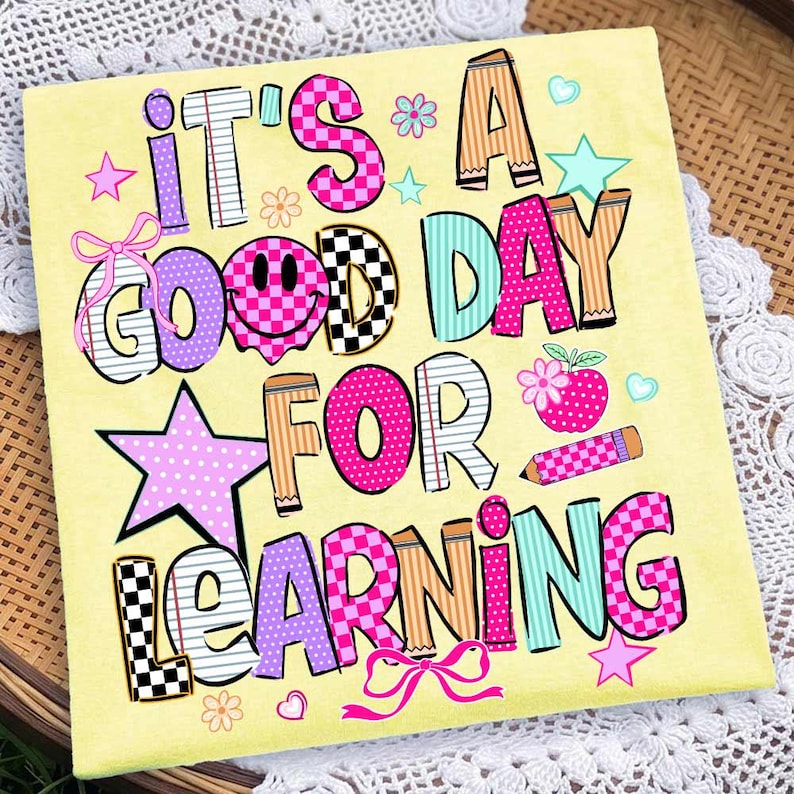 May include: A light yellow t-shirt featuring the colorful text IT'S A GOOD DAY FOR LEARNING. The design includes a pink smiley face, a red apple, a pencil, stars, flowers, and bows in various patterns like checkers, stripes, and polka dots.