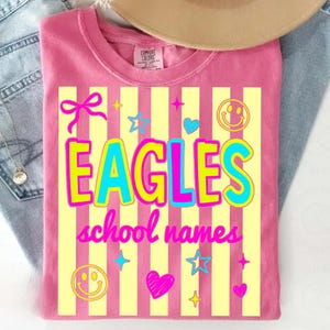 Puede incluir: Camiseta rosa con la palabra "EAGLES" en un dise&ntilde;o en capas de amarillo y azul, sobre rayas verticales amarillas. La camiseta presenta gr&aacute;ficos adicionales como estrellas, corazones y caras sonrientes, con el texto "school names" debajo.
