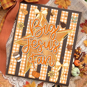 May include: A rectangular sign featuring a background of alternating orange gingham and dark brown stripes. A large gold glitter star sits in the center with the text Big Jesus Fan in white script. Accents include bows, leaves, acorns, and a coffee cup.