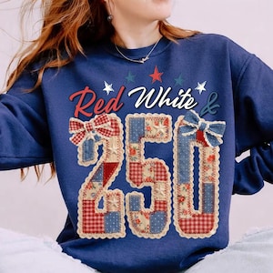 Op de afbeelding: Marineblauwe sweatshirt met de tekst "Red White & 250". De cijfers zijn gemaakt van patchworkstof met rode, witte en blauwe patronen, versierd met strikken. Sterren en strepen maken de feestelijke, patriottische look compleet.