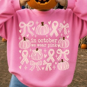 May include: A bright pink sweatshirt featuring a white graphic print of pumpkins, ribbons, and hearts. The text in the center reads, in october we wear pink. The design includes various styles of bows and pumpkins arranged in a grid-like pattern.