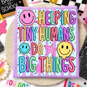 May include: A light purple fabric square featuring a repeating vertical stripe pattern and black polka dots. The design includes the text "HELPING TINY HUMANS DO BIG THINGS" in colorful, patterned block letters, alongside three smiley faces and stars.