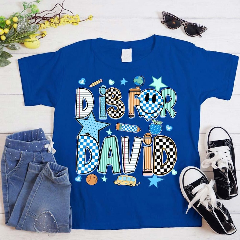 May include: A royal blue short-sleeve t-shirt featuring the text D IS FOR DAVID in a multi-patterned graphic design. The letters incorporate school-themed motifs including checkered patterns, lined paper textures, a smiley face, and a yellow bus.