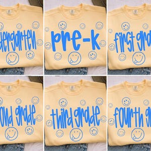May include: A light yellow crewneck sweatshirt featuring blue text and various blue smiley face icons. The text options include kindergarten, pre-k, first grade, second grade, third grade, and fourth grade, all printed in a casual, handwritten font.