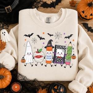 Pode incluir: Uma camisola de gola redonda cor de creme com um gr&aacute;fico colorido de cinco artigos escolares antropom&oacute;rficos vestidos com fatos de Halloween, incluindo um fantasma, uma ma&ccedil;&atilde; m&uacute;mia, uma cola em barra, um caderno vampiro e um l&aacute;pis.