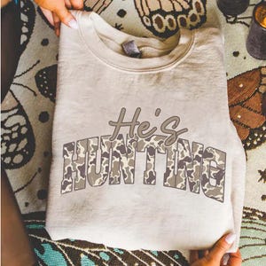 May include: A beige sweatshirt with the text "He's HUNTING" in a camouflage pattern. The sweatshirt is folded and held by hands, showcasing the design. The text is in a brown and gray color scheme.