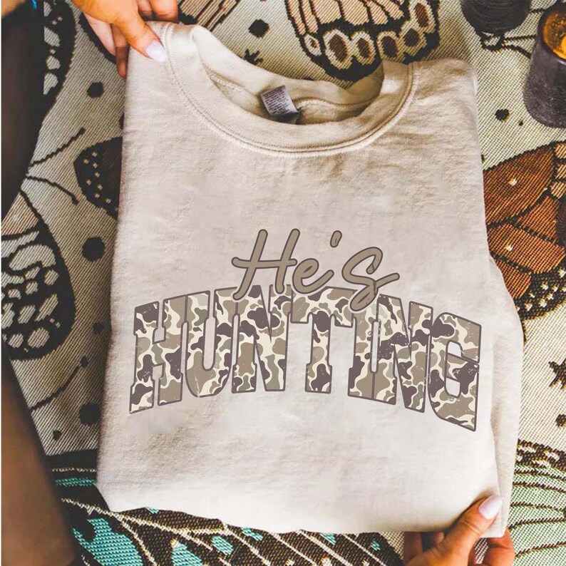 May include: A beige sweatshirt with the text "He's HUNTING" in a camouflage pattern. The sweatshirt is folded and held by hands, showcasing the design. The text is in a brown and gray color scheme.