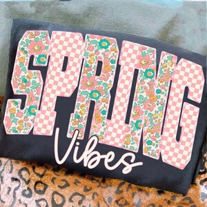 May include: Black t-shirt with the word "SPRING" in large, pink checkered letters with floral accents. The word "Vibes" is written in cursive below the word "SPRING".