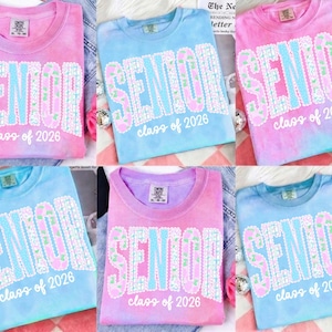 May include: Tie-dye t-shirts in shades of pink, blue, and purple. Each shirt features the word "SENIOR" in large, floral-patterned letters, with "class of 2026" below. The shirts are casual and suitable for graduation.