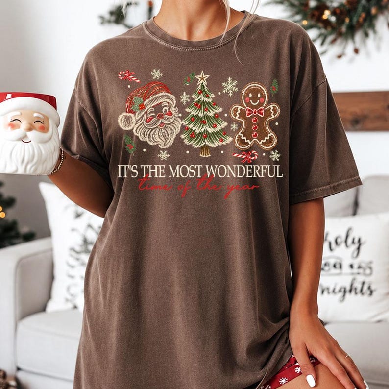 K&ouml;nnte beinhalten: Ein braunes Kurzarm-T-Shirt mit einem Grafikdruck von Weihnachtsmann, einem geschm&uuml;ckten Weihnachtsbaum und einem Lebkuchenmann. Der Text lautet IT'S THE MOST WONDERFUL time of the year in wei&szlig;er und roter Schrift mit kleinen Schneeflocken.