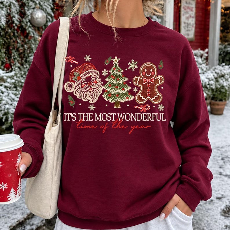 K&ouml;nnte beinhalten: Ein kastanienbraunes Sweatshirt mit Rundhalsausschnitt und einem Grafikdruck, der ein Weihnachtsmann-Gesicht, einen geschm&uuml;ckten Weihnachtsbaum und einen Lebkuchenmann zeigt. Der Text lautet "IT'S THE MOST WONDERFUL time of the year" in wei&szlig;er und roter Schrift.