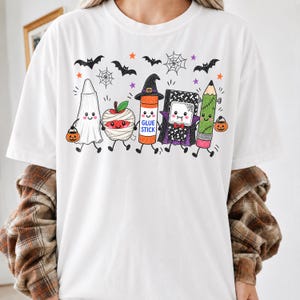 Pode incluir: Uma t-shirt branca com um gr&aacute;fico de cinco materiais escolares antropom&oacute;rficos vestidos para o Halloween: um fantasma, uma ma&ccedil;&atilde; m&uacute;mia, uma cola em bast&atilde;o com chap&eacute;u de bruxa, um caderno vampiro e um l&aacute;pis Frankenstein.