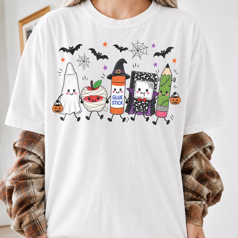 Pode incluir: Uma t-shirt branca com um gr&aacute;fico de cinco materiais escolares antropom&oacute;rficos vestidos para o Halloween: um fantasma, uma ma&ccedil;&atilde; m&uacute;mia, uma cola em bast&atilde;o com chap&eacute;u de bruxa, um caderno vampiro e um l&aacute;pis Frankenstein.