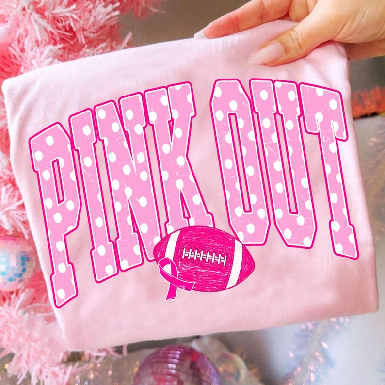 May include: A light pink t-shirt featuring the text PINK OUT in large, bold, white-polka-dotted pink letters. Below the text is a pink American football graphic with a pink awareness ribbon integrated into the design on the left side.