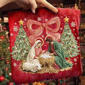 Op de afbeelding: Een rood kerstkussen met een geborduurde kerststal met Maria, Jozef en het kindje Jezus. Twee kerstbomen flankeren de scène, met een grote geruite strik bovenaan. Het kussen wordt door een hand vastgehouden.