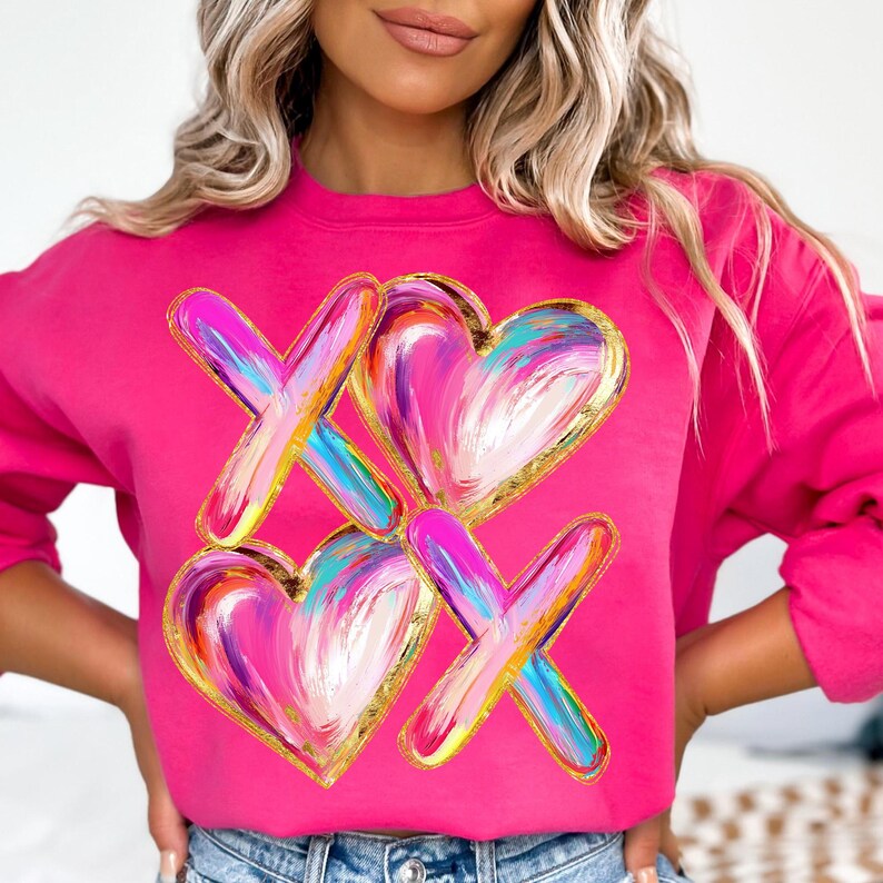 Puede incluir: Sudadera rosa con un gr&aacute;fico colorido de dos corazones y dos X. Los corazones y las X est&aacute;n pintados en un estilo de pinceladas con detalles dorados.