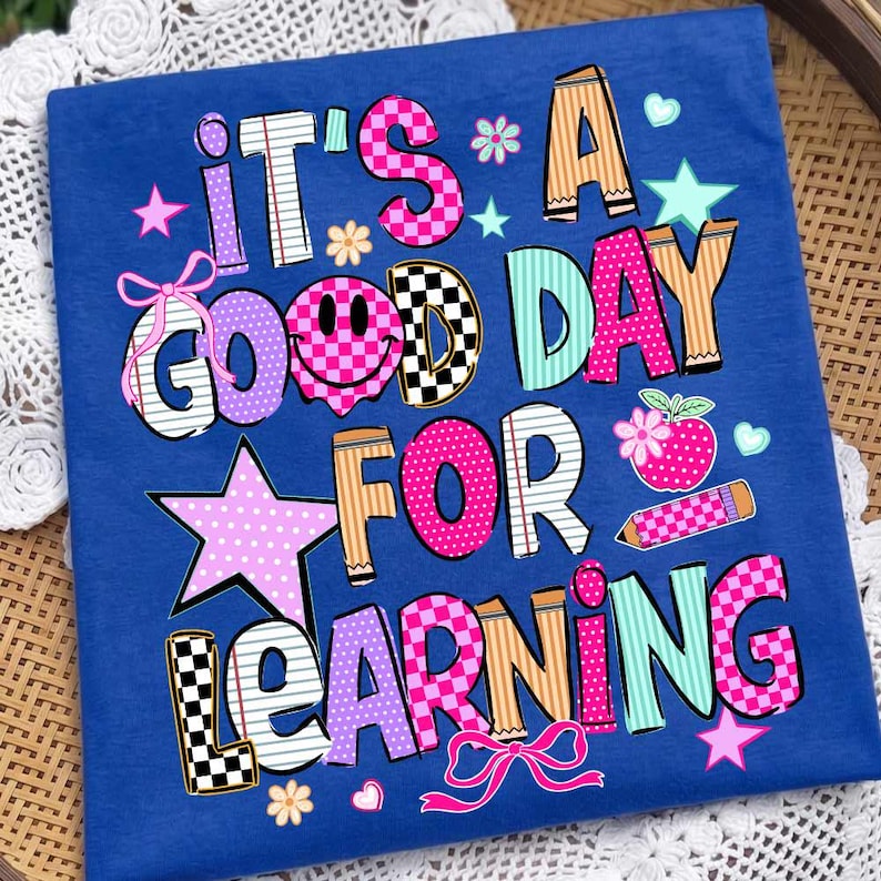 May include: A royal blue t-shirt featuring the text IT'S A GOOD DAY FOR LEARNING in colorful, patterned block letters. The design includes a pink smiley face, stars, flowers, hearts, bows, a pencil, and a red apple with a white flower.