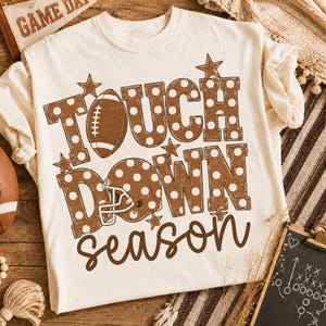 Peut inclure: Un t-shirt couleur crème avec le texte marron TOUCH DOWN à pois blancs. La lettre O de TOUCH est remplacée par un ballon de football américain marron, et le O de DOWN par un casque de football marron. Le mot season est écrit en lettres cursives.