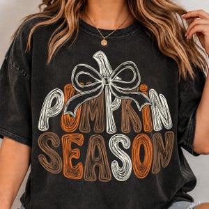 May include: A black mineral-washed short-sleeve t-shirt featuring a graphic of a pumpkin outline topped with a large white bow. The text PUMPKIN SEASON is printed in a stylized, scribbled font in shades of white and orange across the center.