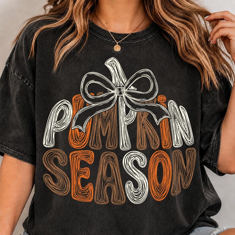 May include: A black mineral-washed short-sleeve t-shirt featuring a graphic of a pumpkin outline topped with a large white bow. The text PUMPKIN SEASON is printed in a stylized, scribbled font in shades of white and orange across the center.