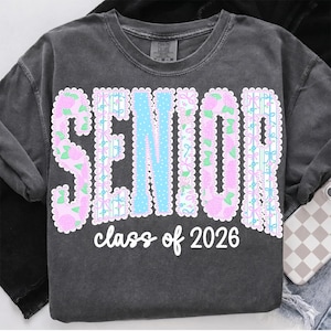May include: Dark gray sweatshirt with the word "SENIOR" in large, decorative letters. The letters are outlined in white and filled with pink floral patterns and light blue accents. The text "class of 2026" is written below in a cursive font.