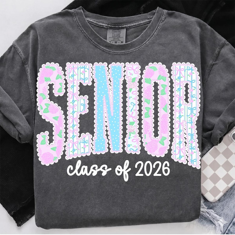 May include: Dark gray sweatshirt with the word "SENIOR" in large, decorative letters. The letters are outlined in white and filled with pink floral patterns and light blue accents. The text "class of 2026" is written below in a cursive font.