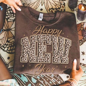 K&ouml;nnte beinhalten: Ein braunes Sweatshirt mit dem Schriftzug "Happy NEW Year" in Gold und Leopardenmuster. Das Sweatshirt wird von H&auml;nden gehalten. Das Design ist festlich und feiert das neue Jahr.