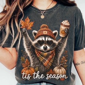 K&ouml;nnte beinhalten: Ein anthrazitfarbenes T-Shirt mit einer Grafik eines Waschb&auml;ren, der eine braune Strickm&uuml;tze, herzf&ouml;rmige Sonnenbrillen und einen karierten Schal tr&auml;gt. Der Waschb&auml;r h&auml;lt ein Ahornblatt und einen Kaffeebecher. Darunter zwei K&uuml;rbisse und der Text "tis the season".