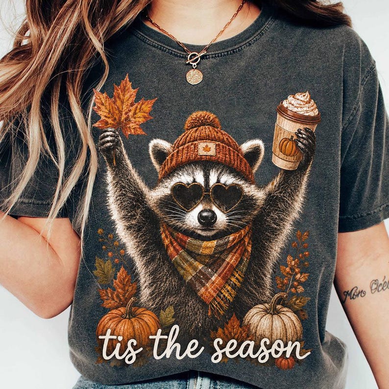 K&ouml;nnte beinhalten: Ein anthrazitfarbenes T-Shirt mit einer Grafik eines Waschb&auml;ren, der eine braune Strickm&uuml;tze, herzf&ouml;rmige Sonnenbrillen und einen karierten Schal tr&auml;gt. Der Waschb&auml;r h&auml;lt ein Ahornblatt und einen Kaffeebecher. Darunter zwei K&uuml;rbisse und der Text "tis the season".