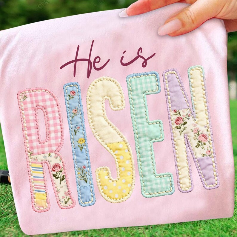 May include: A pink fabric with the words "He is Risen" in a mix of floral and patterned letters. The letters are in pastel colors, including pink, blue, yellow, and green, with a stitched border.
