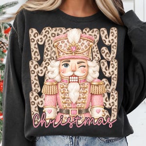 Puede incluir: Una sudadera negra de cuello redondo con el gráfico de un cascanueces guiñando un ojo, vestido con un estampado de leopardo rosa y beige. El texto Merry Merry aparece en estampado de leopardo, con la palabra Christmas en cursiva rosa.