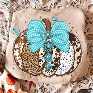 May include: Beige sweatshirt featuring a pumpkin design with a turquoise gingham bow and turquoise jewel accents. The pumpkin has a brown stem and sections with leopard, cowhide, and polka dot patterns. Butterflies and stars are also featured.