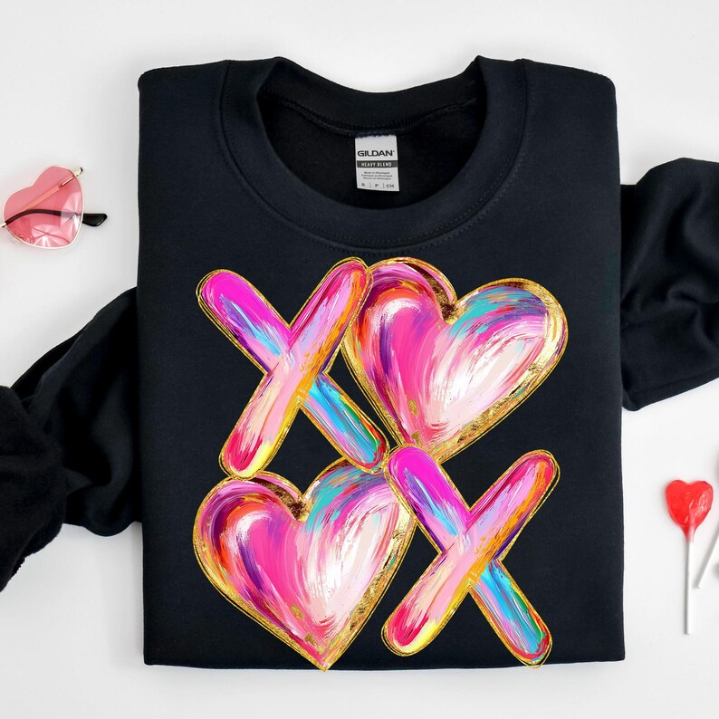 Puede incluir: Sudadera negra con un dise&ntilde;o gr&aacute;fico de dos corazones rosas con detalles dorados y dos X rosas y azules con detalles dorados. El dise&ntilde;o recuerda a un tablero de tres en raya.