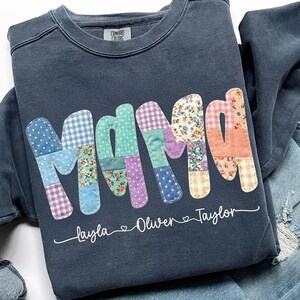 May include: Dark blue sweatshirt with the word "Mama" in patchwork letters. The letters are made of various patterned fabrics, including polka dots and floral prints. The names "Layla", "Oliver", and "Taylor" are written below the word "Mama".