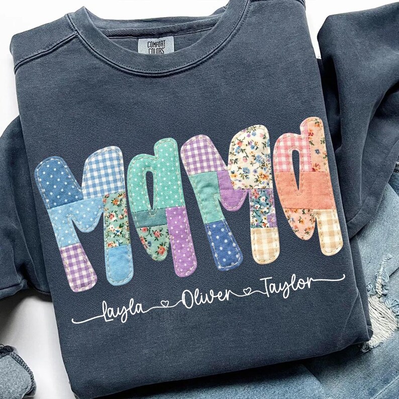May include: Dark blue sweatshirt with the word "Mama" in patchwork letters. The letters are made of various patterned fabrics, including polka dots and floral prints. The names "Layla", "Oliver", and "Taylor" are written below the word "Mama".