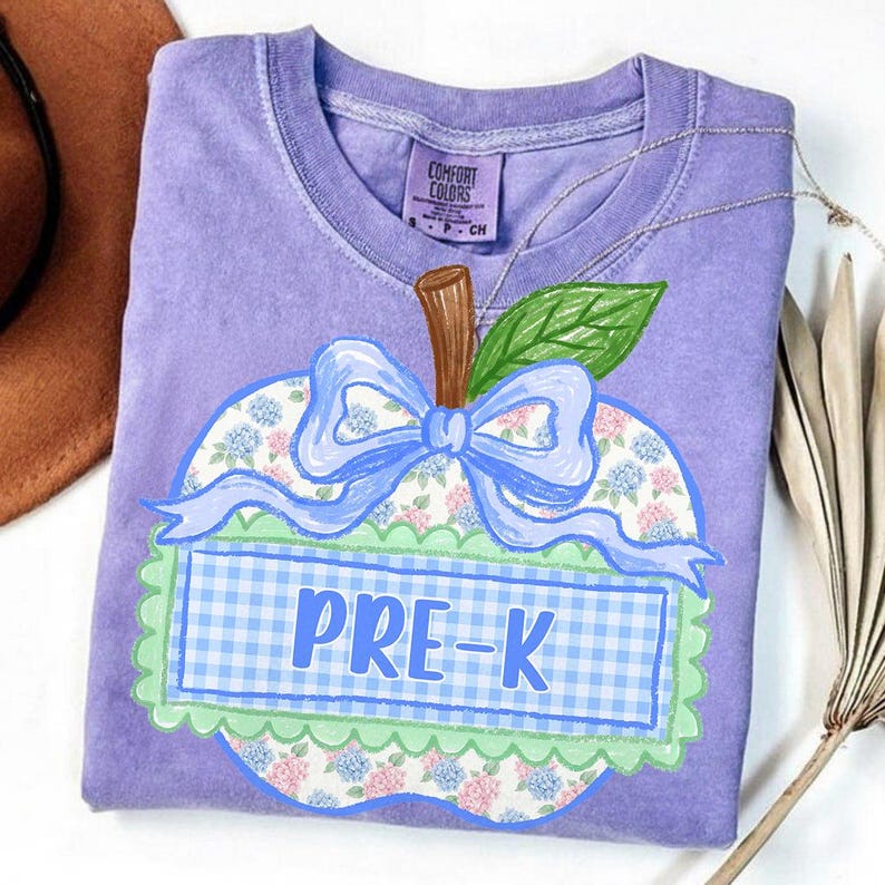 Op de afbeelding: Een paars t-shirt met korte mouwen en een afbeelding van een appel met een bloemenpatroon, een lichtblauwe strik en een groene geschulpte rand. Het midden van de appel bevat een lichtblauw ruitjesrechthoek met de tekst PRE-K in het blauw.