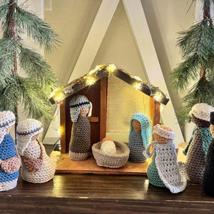 May include: A handcrafted nativity scene featuring crocheted figures in various colors, including blue, beige, and brown. The scene includes a wooden stable with string lights and evergreen branches, creating a festive holiday display.
