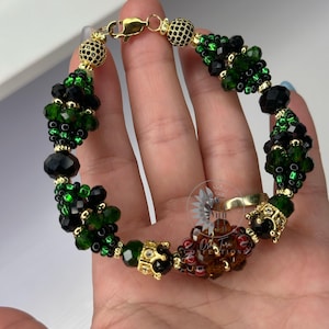 May include: A bracelet with green and black faceted beads, with gold-coloured accents and decorative elements. The bracelet has a gold-coloured clasp and crown-shaped details. The beads are arranged in clusters.