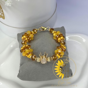 May include: A beaded bracelet with yellow and brown beads, featuring a gold-colored crown charm with clear rhinestones. The bracelet is displayed on a gray pillow. The image also includes a logo with a sunflower and a bee.