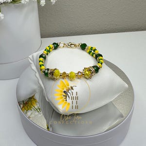 May include: A yellow and green beaded bracelet with gold accents and a clasp. The bracelet is on a white surface with a sunflower design.