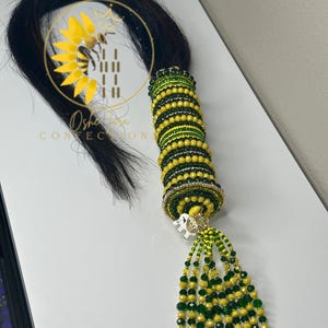 May include: A black hair extension with a decorative beaded wrap in shades of yellow and green. The wrap is adorned with a small white elephant charm and dangling beaded tassels. The logo "Osholara Confections" is visible.