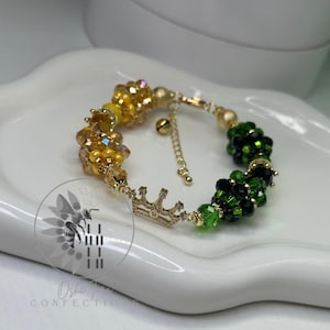 May include: A gold and green beaded bracelet with a crown charm. The bracelet has clusters of beads and a gold chain with a small bell. The bracelet is displayed on a white, curved surface. The beads are arranged in clusters, creating a textured appearance.