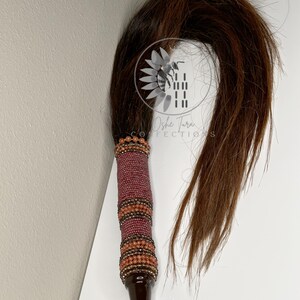 May include: A brown horsehair tail whip with a beaded handle. The handle is made of brown, orange, and pink beads. The tail is long and flowing.