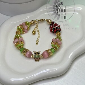 May include: A gold bracelet with pink and green floral beads and an owl charm. The bracelet has a lobster clasp and an extender chain. The bracelet is displayed on a white, wavy surface. The bracelet is approximately 18 cm in length.