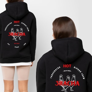 May include: Black hooded sweatshirt with a graphic on the back featuring the text "3RACHA" in red, along with rooster illustrations and additional text. The sweatshirt has a white hem and is worn with beige cycling shorts.