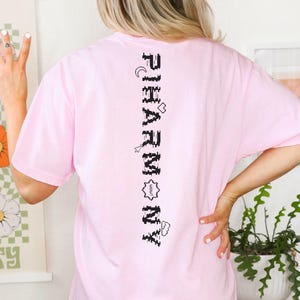 May include: A pale pink t-shirt featuring the word "PHARMONY" printed vertically in black, accompanied by small icons. The t-shirt is a relaxed fit, ideal for casual wear.