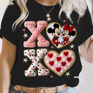 May include: Black t-shirt featuring a Valentine's Day design. The design includes pink knitted "X"s, a heart with Mickey and Minnie Mouse, and a heart filled with red and white hearts. Gold stars and hearts are scattered around the design.