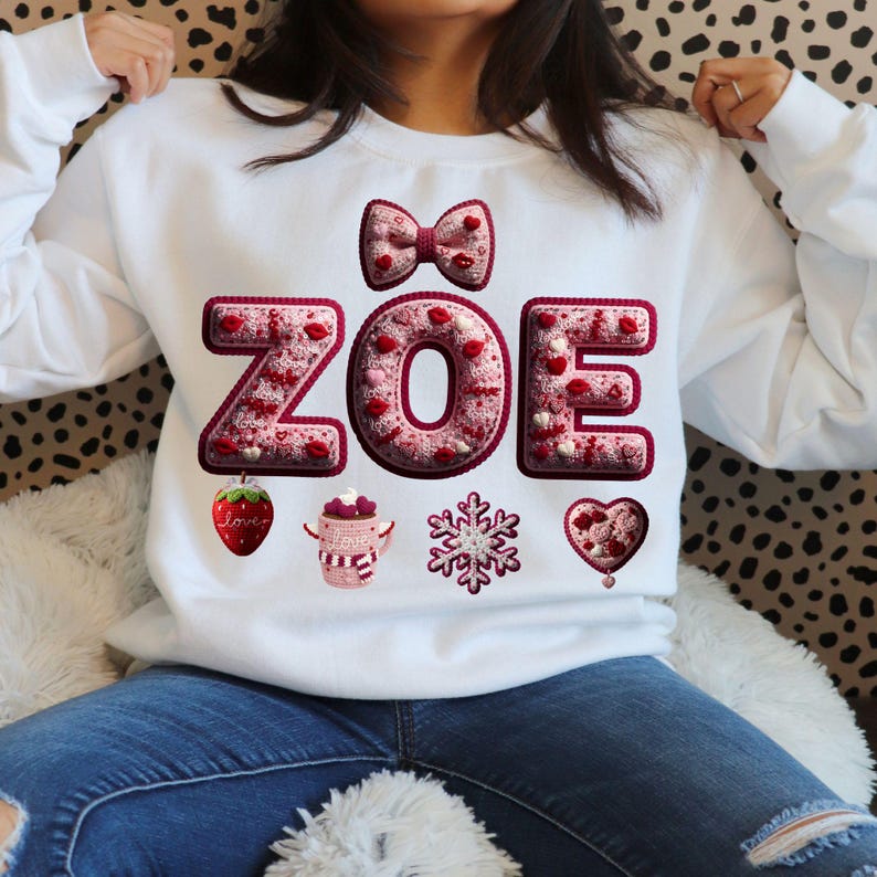May include: White sweatshirt with the name "ZOE" in a decorative font, featuring a bow, hearts, and other embellishments. The design includes a strawberry, a mug, a snowflake, and a heart. The sweatshirt is a casual, everyday garment.