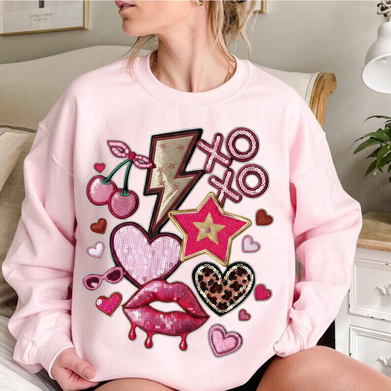 May include: A light pink sweatshirt featuring various patches, including cherries, a lightning bolt, a star, hearts, sunglasses, lips, and the letters "XO". The patches are in shades of pink, red, and gold.
